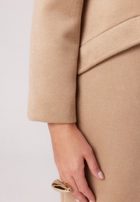 Beżowy wełniany płaszcz z rękawem, charakteryzujący się eleganckim, dopasowanym krojem, z przeszywaną kieszenią i ręką ozdobioną złotym pierścionkiem.