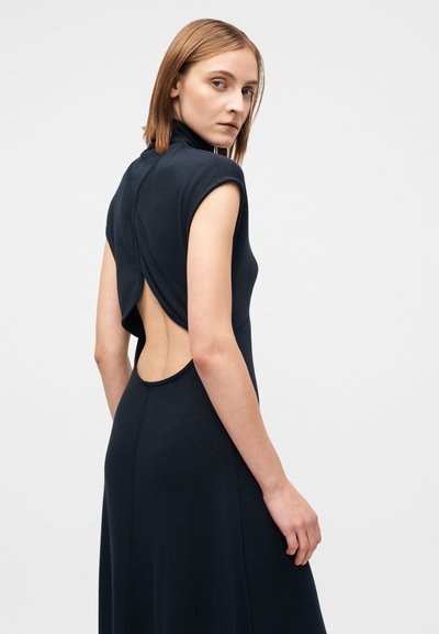 Ujjatlan tengerészkék ruha magas nyakkal, nyitott hátrésszel és redőzött részletekkel. Puha anyagból készült, testhez simuló sziluettel és lebegő szoknyával.