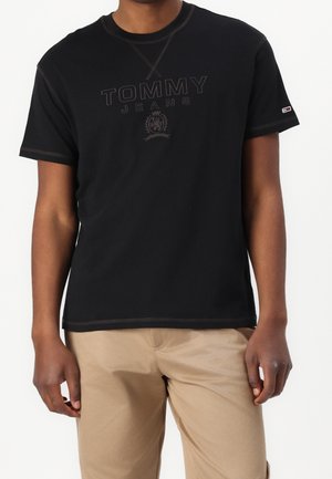 Osoba nosząca czarną krótką koszulkę Tommy Jeans z logo oraz beżowe spodnie, stojąca na jednolitym białym tle.
