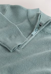 Pull en maille bleu clair avec col à fermeture éclair quart et poignets côtelés, posé à plat sur une surface blanche.