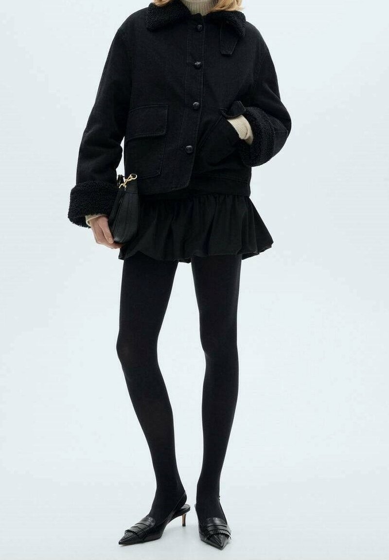 Personne portant une veste en peau lainée noire, une jupe noire à volants, des collants noirs, des escarpins noirs à bout pointu avec un petit talon et tenant un petit sac noir.