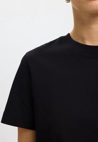 Personne portant un t-shirt noir uni à col rond et manches courtes sur un fond blanc avec une petite boucle d'oreille argentée visible.