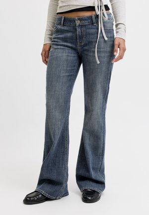 Femme portant un jean taille haute bleu délavé à jambes larges, des chaussures noires et un top court à manches longues léger avec des cordons.