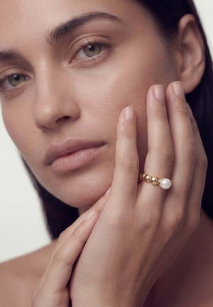 Femeie cu piele deschisă și ochi verzi care își atinge ușor fața, purtând un inel de aur cu o perlă mare pe degetul mijlociu.