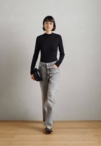 Melna ilga piedurkne augšdaļa, gaiši pelēki augstās jostasvietas džinsi, melna rokassoma un rakstainas čības, stāvot uz gaišas koka grīdas.
