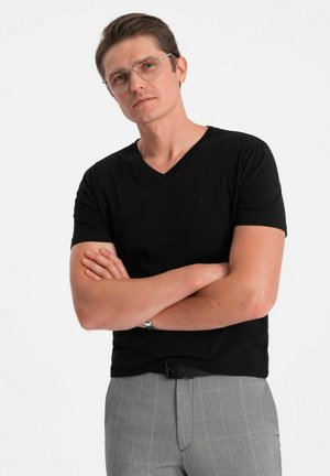 Camiseta negra con cuello en V hecha de tela suave, combinada con pantalones de cuadros grises. El modelo tiene el cabello corto y lleva gafas con montura plateada.