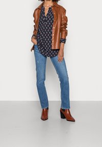 Braune Lederjacke über einem dunkelblumigen Hemd mit Knöpfen, kombiniert mit hellblauen Jeans und braunen Absatzstiefeln. Schlichter Hintergrund.