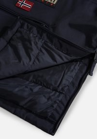 Veste bleu marine foncé partiellement zippée dévoilant une doublure intérieure matelassée et lisse ainsi qu'un patch drapeau norvégien cousu près du col.