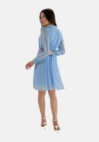 Abito blu chiaro con maniche lunghe, realizzato in materiale chiffon leggero, con cintura in vita e una gonna fluida. Abbinato a tacchi bianchi a strappo.