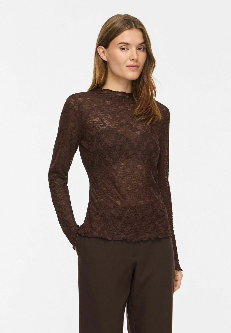 Femme aux cheveux bruns portant un haut ajusté à manches longues en dentelle florale marron foncé et un pantalon marron foncé, debout avec une main dans la poche.