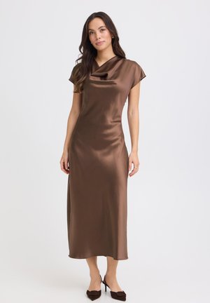 Naine, kes kannab pruunist satiinist midi-pikkusega kleiti kapuutsvarrukatega ja drapeeritud kaelusega, kombineeritud tumedate pruuni varbaga kontsakingadega, seisab valge tausta ees.