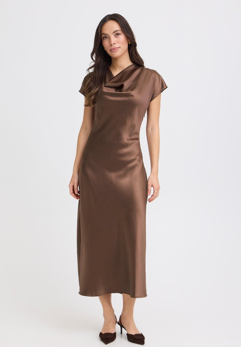 Femme portant une robe midi en satin marron avec des manches courtes et un col drapé, associée à des escarpins pointus marron foncé, debout devant un fond blanc.