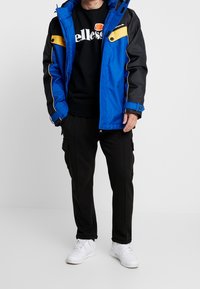 Hombre con una chaqueta azul y negra, sudadera negra de ellesse, pantalones negros y zapatillas blancas, de pie contra un fondo blanco liso.