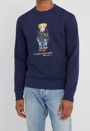Sweat-shirt bleu marine présentant un ours vêtu d'un blazer, d'un pull, d'un jean et de bottes avec le texte « Polo Bear by Ralph Lauren » en dessous.