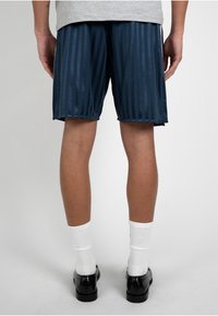 Personne portant un short rayé bleu marine, des chaussettes blanches, des chaussures habillées noires et une chemise grise, debout, dos à un fond uni.