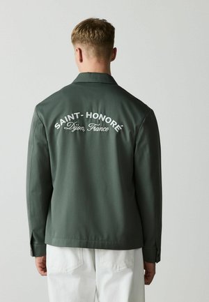 Man wearing dark green jacket with "SAINT-HONORÉ Dijon, France" text on back, white pants, standing facing a plain gray background.