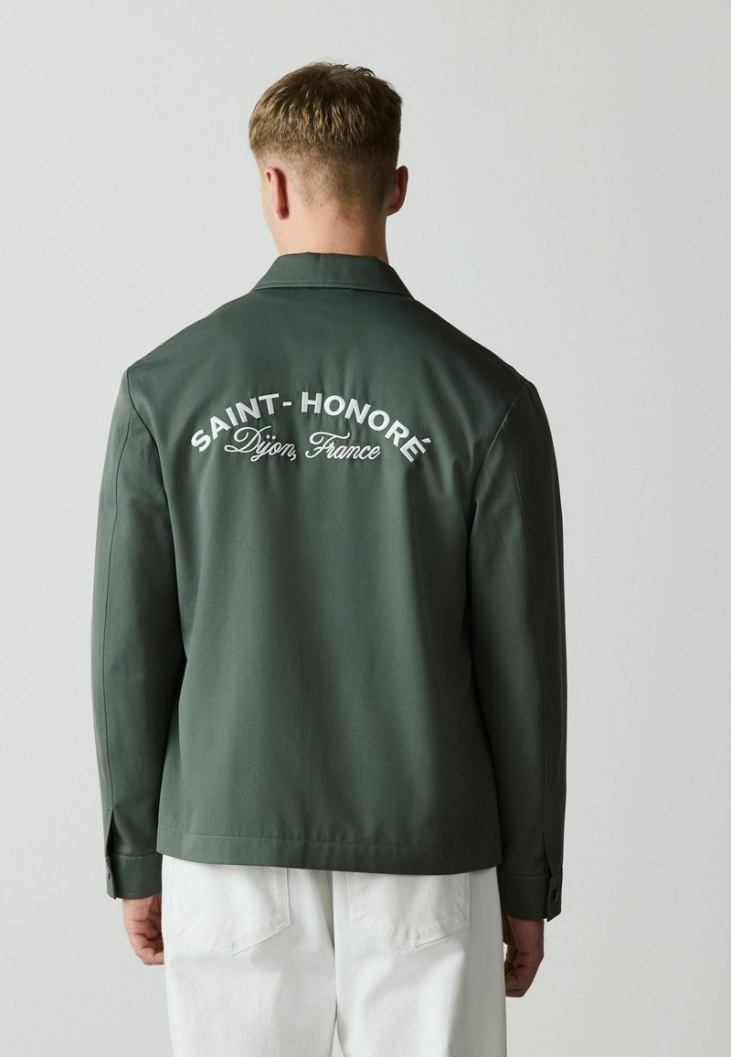 Homme portant une veste vert foncé avec le texte « SAINT-HONORÉ Dijon, France » au dos, pantalon blanc, debout face à un fond gris uni.