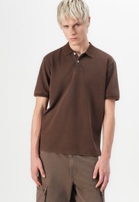Brown polo shirt made of soft cotton, featuring a classic collar, two-button placket, and short sleeves. Paired with cargo pants.