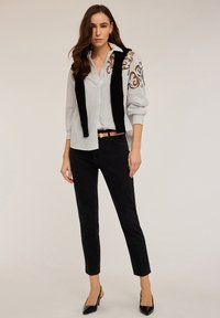 Camisa de botones blanca a rayas con acentos bordados, combinada con pantalones ajustados negros y zapatos de tacón negros, con un suéter negro drapeado.