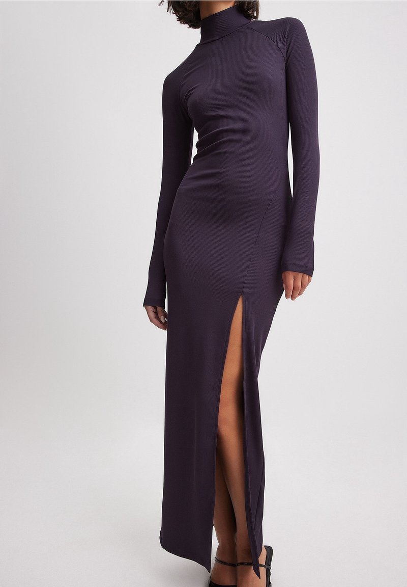 Femme portant une robe longue moulante à manches longues, col montant, de couleur violet foncé, avec une fente haute sur un côté, associée à des talons noirs à brides.