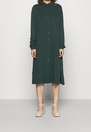 Femme portant une robe midi vert foncé à manches longues, boutonnée avec un col, et des chaussures Mary Jane beiges à talon bloc.