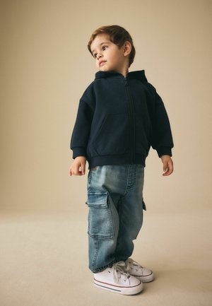 Laivastonsininen vetoketjullinen huppari, vaaleansiniset farkkukangassortit taskuilla ja valkoiset lenkkarit punaisilla yksityiskohdilla. Sileä kangaspinta ja rento istuvuus.