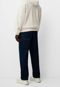Persona vestida con una sudadera color crema, jeans de pierna ancha azul oscuro y zapatillas blancas, de pie frente a un fondo blanco liso.