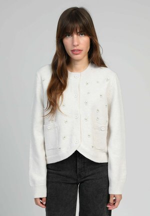 Femme aux longs cheveux bruns portant un cardigan blanc à boutons avec de petits détails en perles et un pantalon noir devant un fond uni.