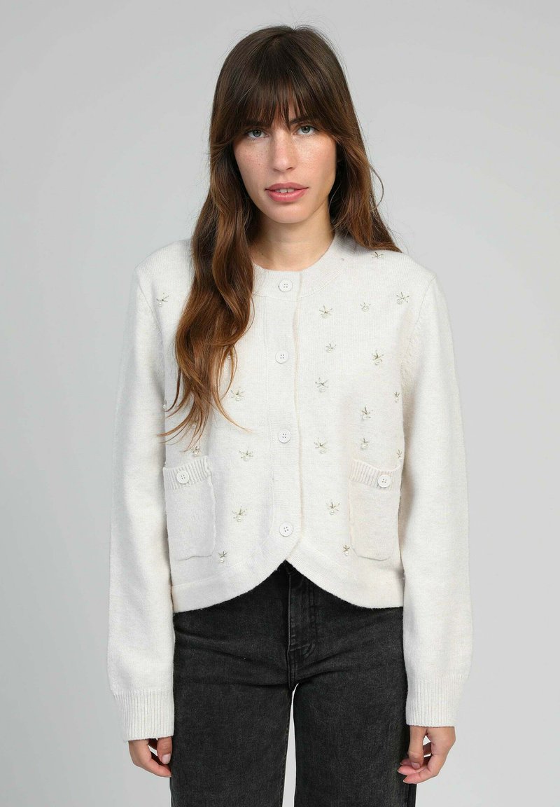 Femme aux longs cheveux bruns portant un cardigan blanc à boutons avec de petits détails en perles et un pantalon noir devant un fond uni.