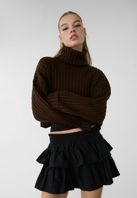 Brun ribbad turtlenecktröja med överdimensionerade ärmar, tillsammans med en svart, lager på lager-rufflad kjol med elastisk midja.
