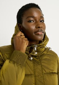 Doudoune verte olive avec capuche, présentant des poignets côtelés et une fermeture éclair à l'avant. Accents en matériel argenté et cordon de serrage pour un look ajusté.