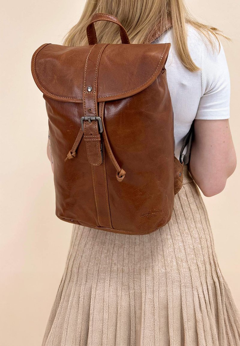 Zaino in pelle marrone con patta curva, spallacci regolabili e componenti in metallo. Presenta una texture liscia e una leggera decorazione in rilievo.