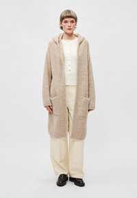 Cardigan en tricot beige, oversize, avec une capuche, deux poches avant et une finition texturée, porté sur un haut button-up de couleur claire.