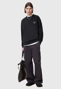 Felpa nera con design grafico a forma di cuore, abbinata a pantaloni cargo neri larghi e scarpe slip-on nere, mentre tiene una borsa nera.