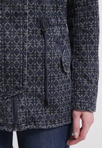 Blå jacka med mönster i geometriska former, med dragkedja, dragsko i midjan och en sidoficka med klaff. Tyget verkar vara texturerat.