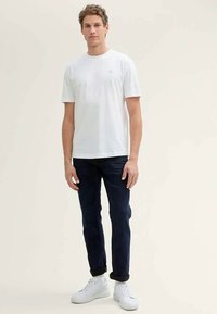 Joven de pie, vistiendo una camiseta blanca sencilla, jeans oscuros arremangados y zapatillas blancas sobre un fondo claro.