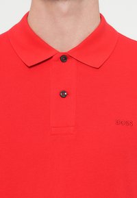 Czerwona koszulka polo z fakturowanej tkaniny, z klasycznym kołnierzykiem, dwoma czarnymi guzikami oraz haftowanym logo "BOSS" na piersi.