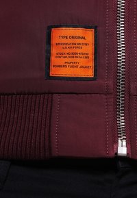 Patch rectangulaire orange avec texte noir sur une veste bomber bordeaux près de la fermeture éclair argentée et du poignet côtelé.