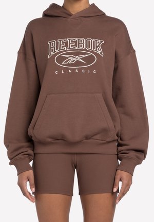 Personne portant un sweat à capuche oversize marron Reebok Classic avec poche avant et shorts biker côtelés assortis.