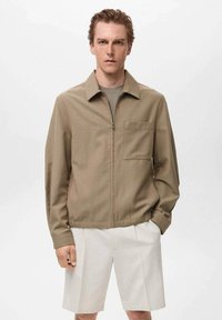 Giacca khaki con zip, colletto, taschino sul petto e taglio dritto. Indossata sopra un maglione grigio a trama e pantaloni corti bianchi.