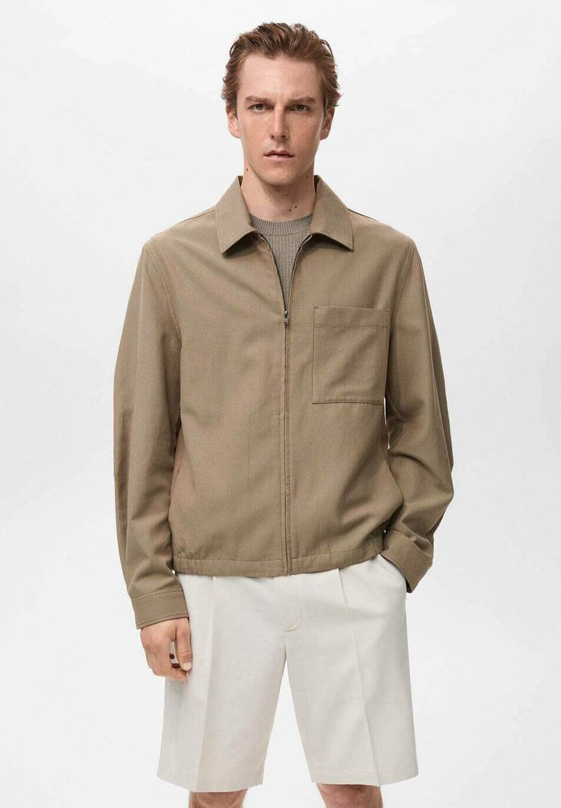 Giacca khaki con zip, colletto, taschino sul petto e taglio dritto. Indossata sopra un maglione grigio a trama e pantaloni corti bianchi.