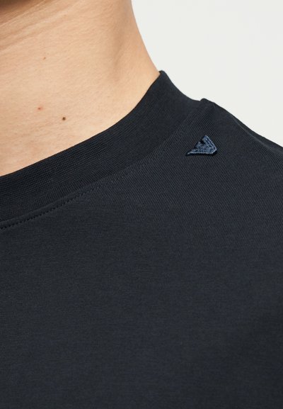 Κοντινή λήψη του λαιμού και του ώμου ενός ατόμου που φοράει μαύρο μπλουζάκι με στρογγυλή λαιμόκοψη και μικρό σκούρο κεντημένο λογότυπο κοντά στον γιακά.