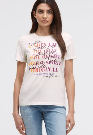 Short-sleeve beige t-shirt with multicolored text prints, featuring various fonts. Paired with light blue denim jeans.
