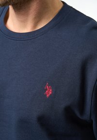 Marineblauwe katoenen T-shirt met ronde hals, voorzien van een klein rood geborduurd logo op de linkerkant van de borst. Effen structuur en een getailleerd ontwerp.