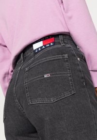 Svarta denimjeans med klassiska bakfickor, pryds av en blå och röd märkespatch. Matchas med en ljuslila sweatshirt.