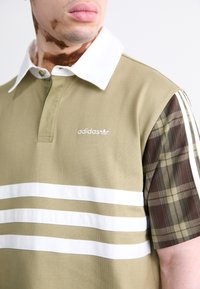 Olivnozeleni polo majica z belimi črtami, kontrastnimi karirastimi rokavi, belim ovratnikom in Adidasovim logotipom na prsih. Tekstuirana bombažna tkanina.