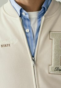 Giacca beige con zip e colletto a coste, indossata sopra una camicia a righe blu e bianche. Presenta testo ricamato e un design con lettera testurizzato.