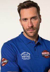 Modrá polo košile s designem inspirovaným klasickými automobily. Obsahuje vyšívané loga na rukávech a límci, vyrobená z hladké bavlněné směsi.