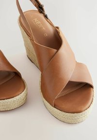 Brown leather wedge sandals with crisscross straps, jute-wrapped platform, and adjustable ankle strap. Smooth texture with subtle stitching.