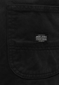 Μαύρη τσέπη από ύφασμα με ραμμένη ορθογώνια ετικέτα που γράφει "JACK & JONES Cotton Dry Goods Div. Established 1990."